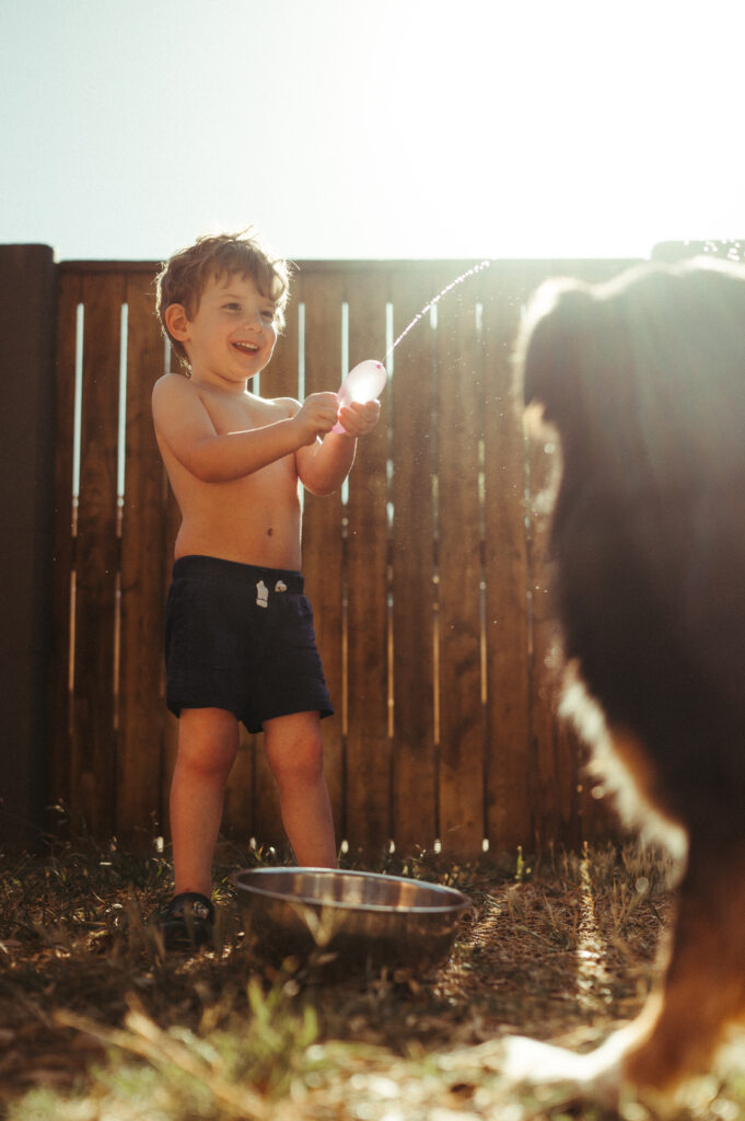 Young boy squirting a water balloon at his dog and giggling. 